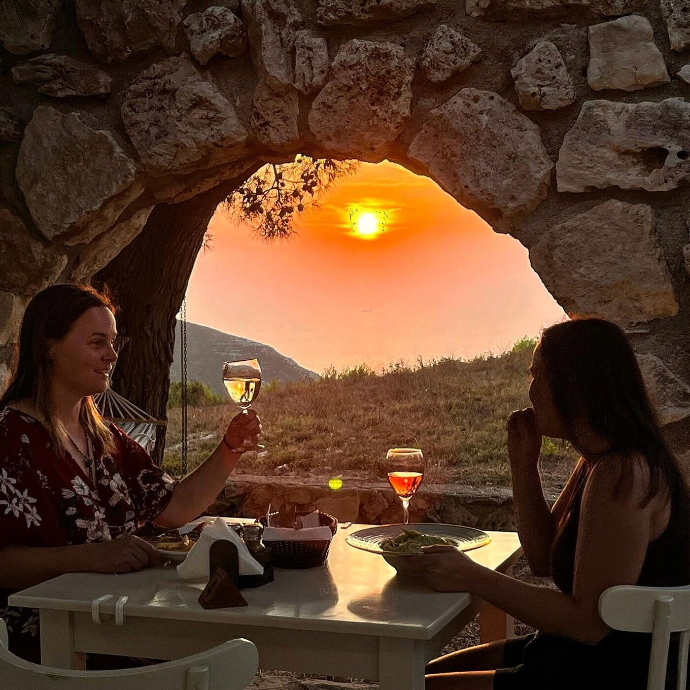 Two guests toasting wine glasses under a stone archway at sunset